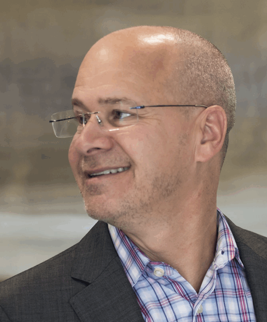 Smiling bald man wearing glasses, a plaid button-up shirt, and a dark blazer, looking to the side against a blurred background.